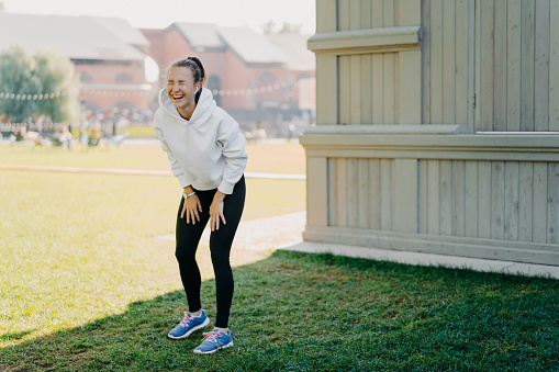 Cheerful sporty woman laughs happily has fun while doing sport exercises listens music via earphones wears white hoodie sportshoes poses outdoors on green grass. People emotions active lifestyle