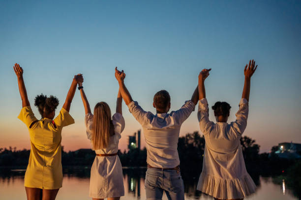 Rear view of cheerful young friends with arms raised holding hands and standing at lakeshore with clear sky in the background