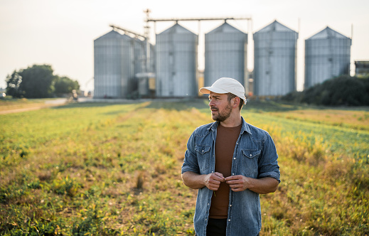 Male farmer standing in agricultural field with silos in background on sunny day