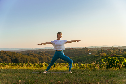 Caucasian young woman with arms outstretched practicing warrior 2 pose on grassy land against clear sky during sunrise