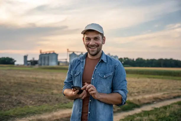 Smiling male farmer text messaging on smart phone while standing in agricultural field against sky