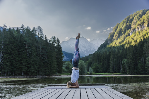 Side view of young female yogi practicing headstand on pier surrounded with lake against pine trees and sky in forest