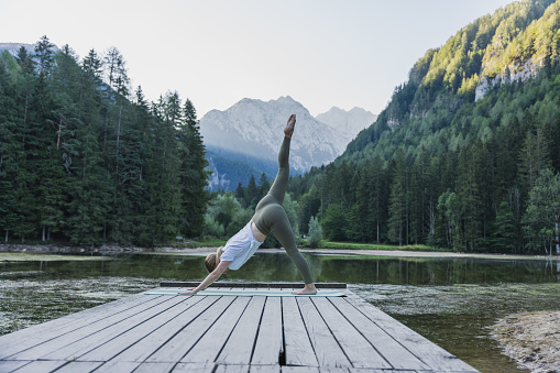 Young woman with leg up practicing downward facing dog position on pier amidst lake against pine trees growing in forest