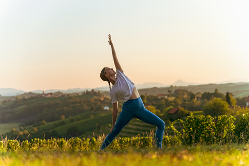 Young female Caucasian yogi practicing viparita virabhadrasana with lush landscape and clear sky in the background during sunrise