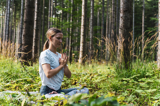 Young female yogi with eyes closed meditating in prayer position amidst green plants against trees in forest during getaway