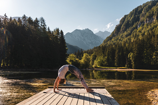 Caucasian young woman practicing upward facing bow pose on pier amidst lake against lush pine trees and sky in forest