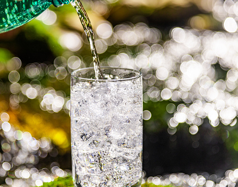 Close-up of clean water filling in drinking glass with ice cubes against defocused river at rainforest during sunny day