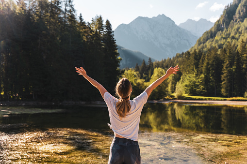Rear view of young woman with arms raised practicing mountain pose by lake against pine trees in forest during sunny day