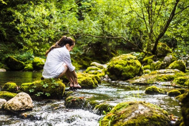 Side view of Caucasian young woman sitting on mossy rock amidst river flowing in lush rainforest during vacation