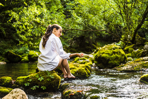 Rear view of carefree young woman playing with water while sitting on mossy rock amidst river flowing in rainforest