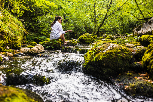 Low angle view of young woman playing with water while sitting on rock at riverbank with lush trees growing in the background at forest