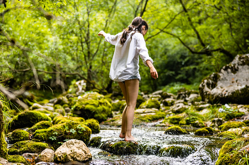 Rear view of playful young woman with arms outstretched standing on rocks amidst flowing river in rainforest during vacation