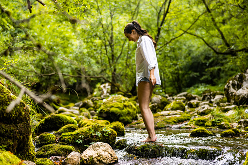 Side view of carefree young woman standing on rock amidst flowing river against trees growing in rainforest during vacation