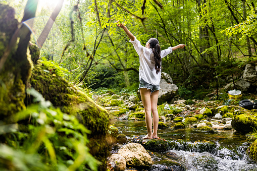 Low angle view of carefree young woman with arms outstretched standing on rock amidst flowing river in lush forest