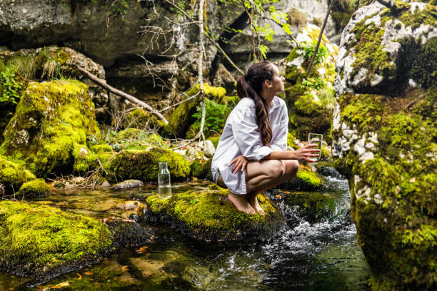 istock Thoughtful woman with drinking glass crouching on rock by river 1451091806