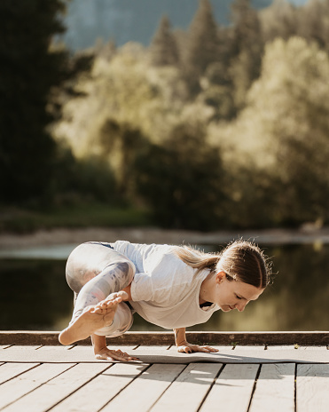 Dedicated Caucasian young female yogi practicing side crow pose on pier against lake and trees in forest during sunset