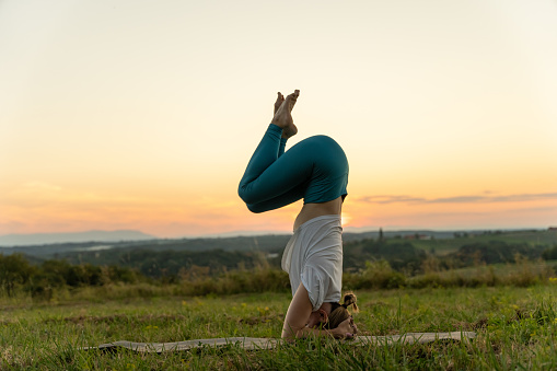 Side view of Caucasian young woman practicing garuda salamba sirsasana on exercise mat over grassy landscape against sky