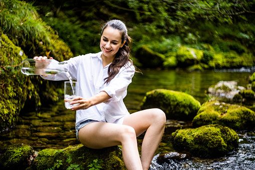 Smiling beautiful young woman pouring clean water in drinking glass while sitting on rock amidst river in rainforest