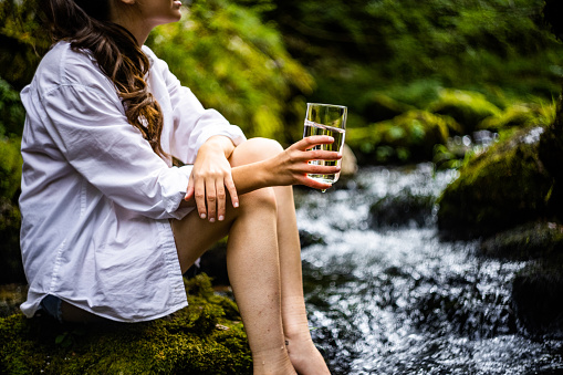 Midsection of young woman holding drinking glass with clean water while sitting on rock by flowing river in forest