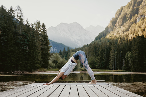 Side view of young female yogi practicing downward facing dog position on pier amidst lake against lush pine trees in forest