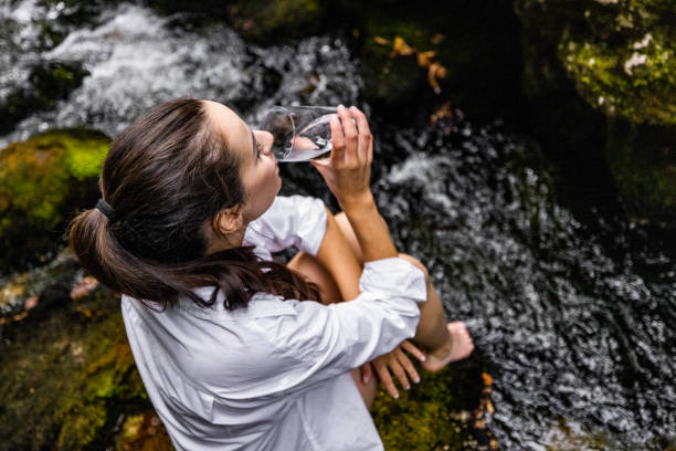 istock Woman drinking water while sitting on rock by river in forest 1451092128