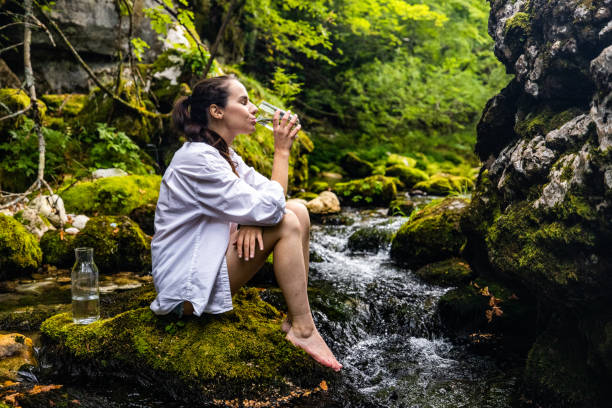 istock Thirsty woman drinking water while sitting on rock by river 1451092150