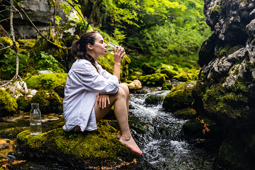 Side view of thirsty beautiful woman drinking water while sitting on rock by flowing river in rainforest during vacation