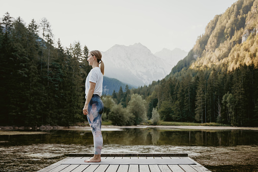 Side view of young woman standing in mountain pose on exercise mat over pier by lake against pine trees at forest