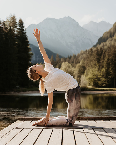 Side view of Caucasian young female yogi with hand raised practicing camel pose on pier by lake in pine forest during sunset