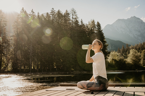 Tired Caucasian young woman drinking water while sitting on pier amidst lake against pine trees in forest at sunset