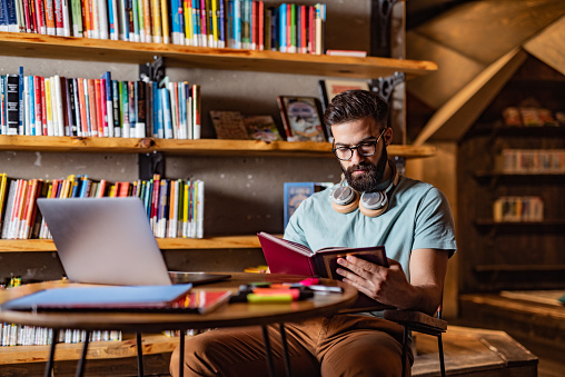 Young male student studying in the library while using a laptop and book