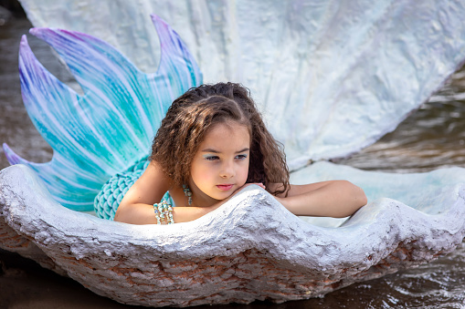 A beautiful little brunette girl in a turquoise mermaid costume lie in a large sea shell, outdoor, looking away. Close up. Copy space