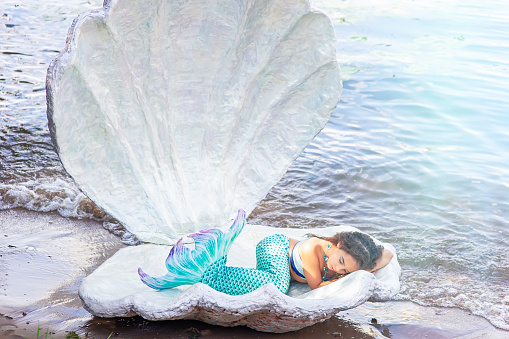 A pretty little brunette girl in a turquoise mermaid costume sleeping in a large sea shell, outdoor. Close up. Copy space