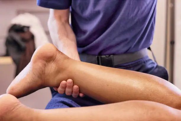 The close-up photograph shows a male massage therapist's hands doing leg massage on a female client on a massage bed inside a clinic.