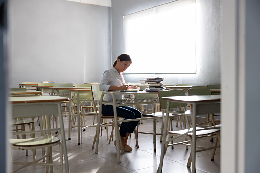 Latin American teacher in a empty classroom at the school grading some tests - education concepts