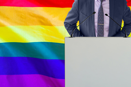 Tribune with microphone and man in suit on LGBT flag background. Businessman and tribune on LGBT flag background. Politician at the podium with microphones background Rainbow flag. Conference LGBT