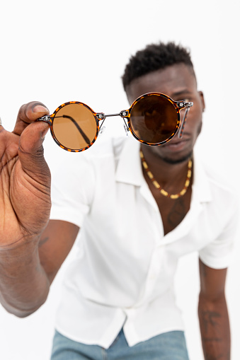 African-American man in white shirt wearing sunglasses against white background. Black male model holding his glasses and showing them to the camera.