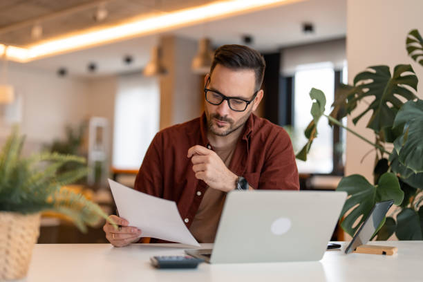 Businessman constantly working stock photo Serious millennial man using laptop sitting at the table in a home office, focused guy in casual clothing looking at the paper, communicating online, writing emails, distantly working or studying on computer at home. small business stock pictures, royalty-free photos & images
