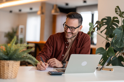 Shot of a young satisfied businessman using a laptop and writing business plan on a paper at his working place. Happy business man working with documents and laptop. Writing down ideas and using laptop.