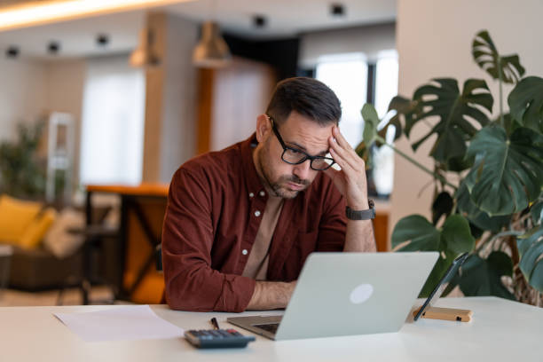 Financial problems Contemplating businessman in the home office feeling frustrated. Full concentration on work. Man working on laptop, counting profit while sitting at the desk at home. head in hands stock pictures, royalty-free photos & images