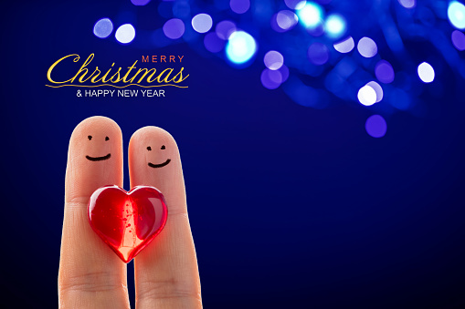 Merry Christmas and Happy New Year text on a blue bokeh lights background. Happy finger couple holding a red heart on the front. Space for copy.
