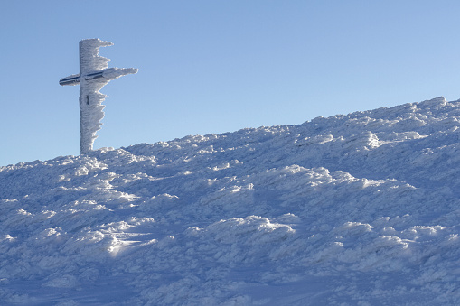Metal iron frozen cross, completely covered by snow and icicles and mountain winter landscape under blue sky