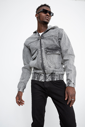 Black male model with sunglasses wears a denim jacket and is in front of a white background.