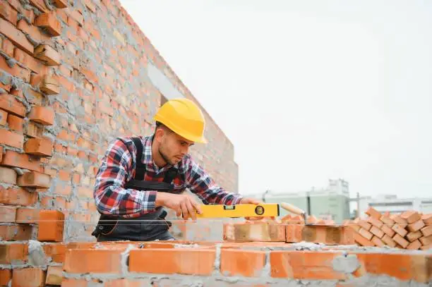 Photo of construction mason worker bricklayer installing red brick with trowel putty knife outdoors.