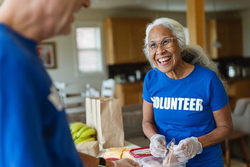 Cheerful mature adult woman of Pacific Islander descent smiles and talks with her husband while working as a volunteer preparing meals for those in need. Charity and relief work concept.