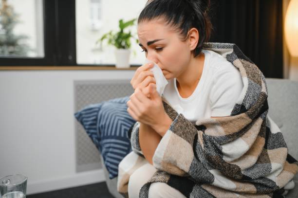 istock Sick woman with a headache sitting on a sofa at home wrapped in grey blanket 1451145629