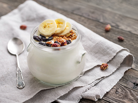 Natural homemade yogurt in a glass jar. Healthy food for breakfast with muesli. Jar with granola and banana slices on linen tablecloth on  wooden table.