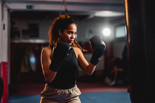Strong young Hispanic female athlete exercising with a punching bag. She is indoors in a gym, wearing sports clothing and boxing gloves.