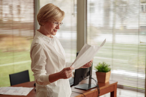 istock Pretty mature woman freelancer works with documents while standing on background of home office 1451155452