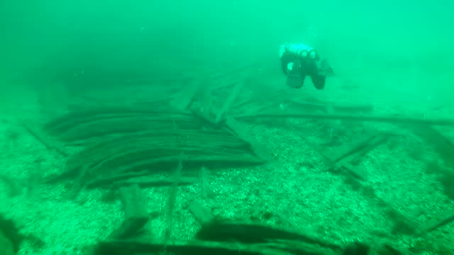 The camera slowly pans the seabed with wooden debris, a diver swims above them. Shipwreck of an 18th century merchant sailing ship.